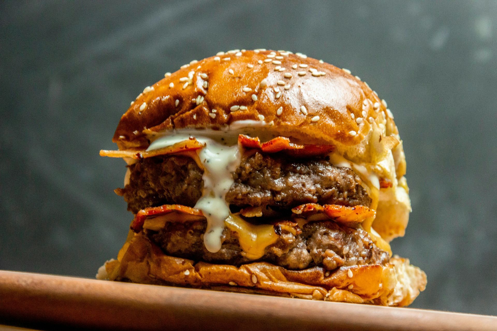 A juicy double cheeseburger with bacon and melted sauce on a toasted sesame bun, set against a dark gray background.