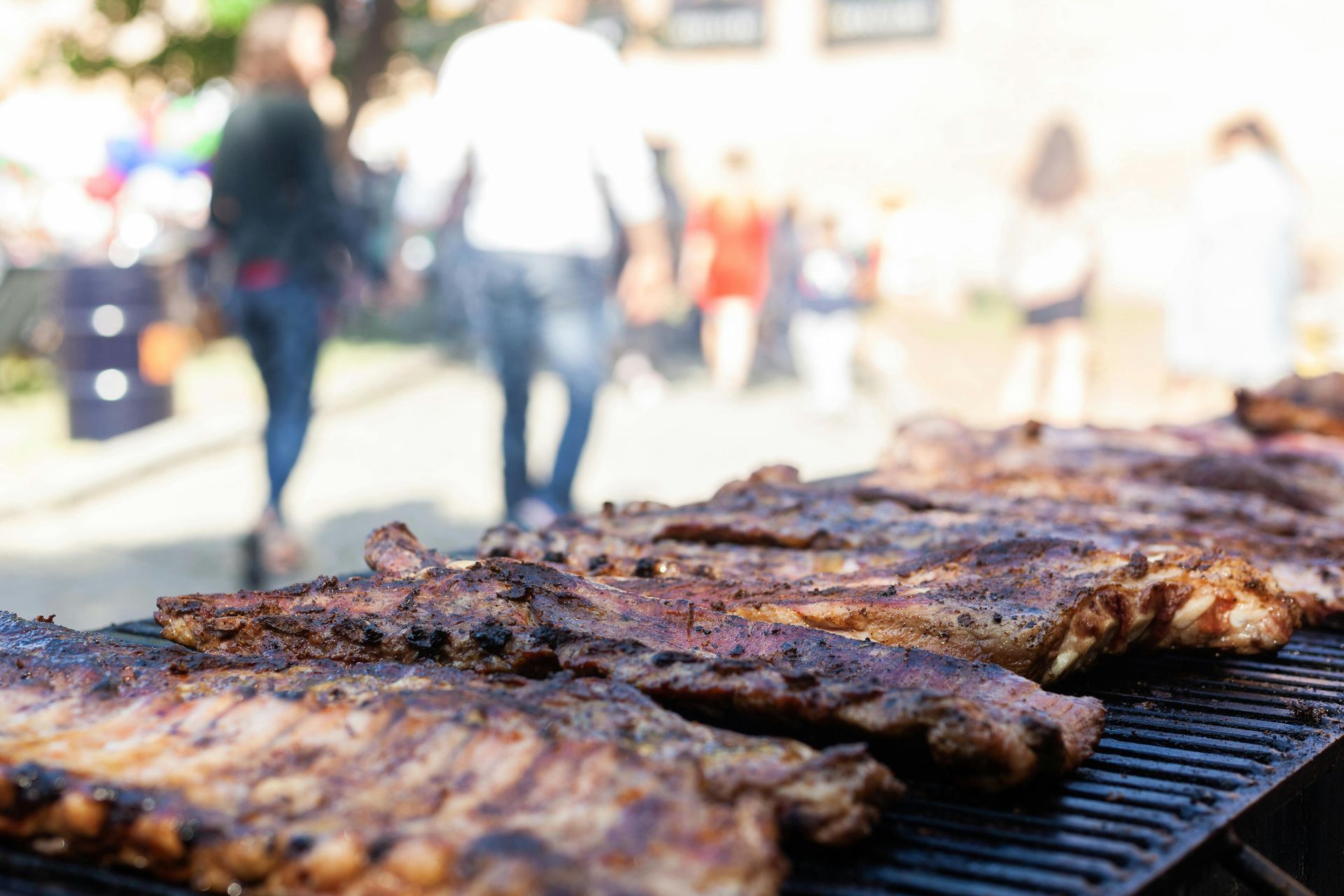 Grilled ribs cooking on a barbecue grill at an outdoor event with blurred people walking in the background.