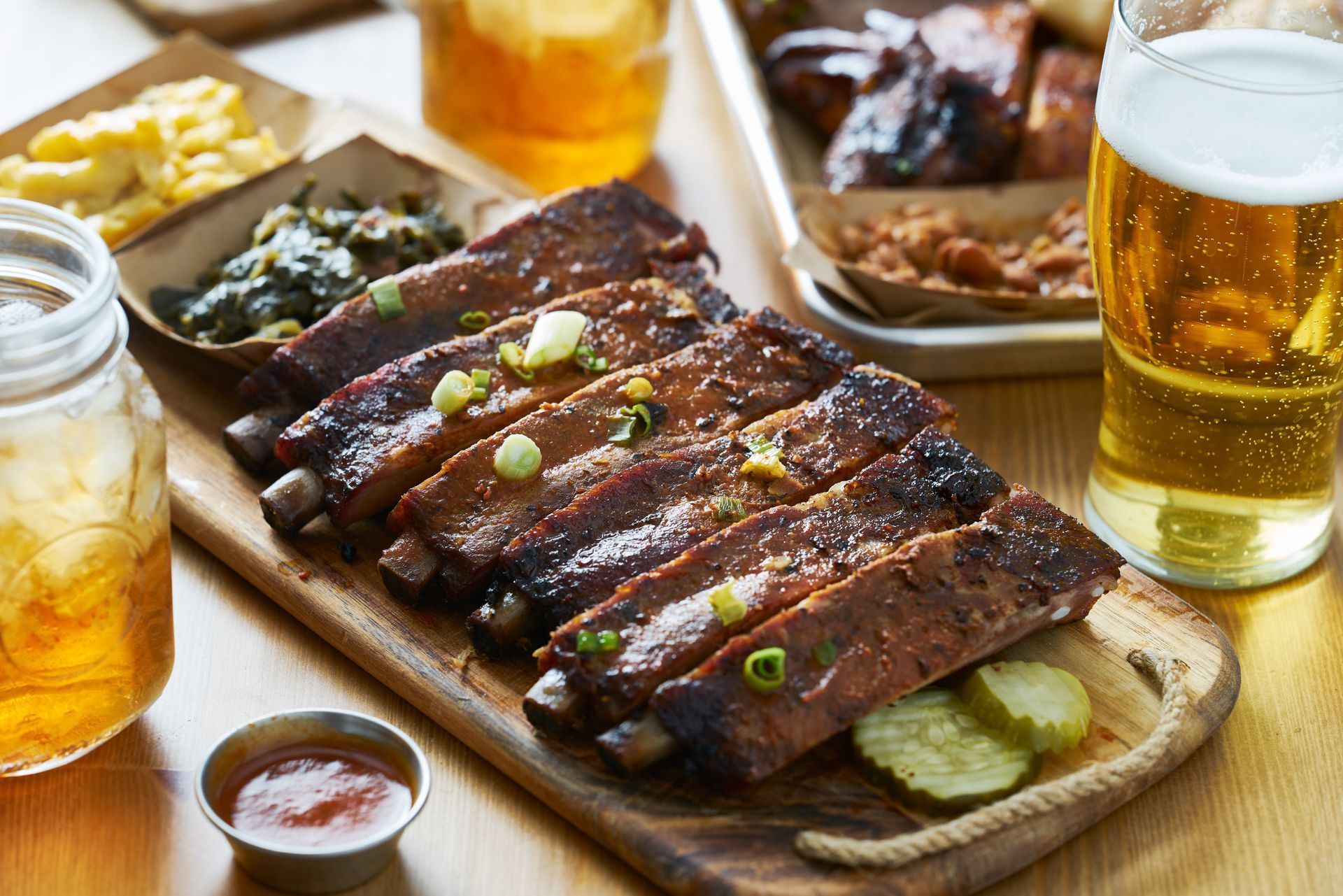 A wooden board features a rack of glazed ribs topped with green onions, next to small bowls of sides, pickles, and a beer.