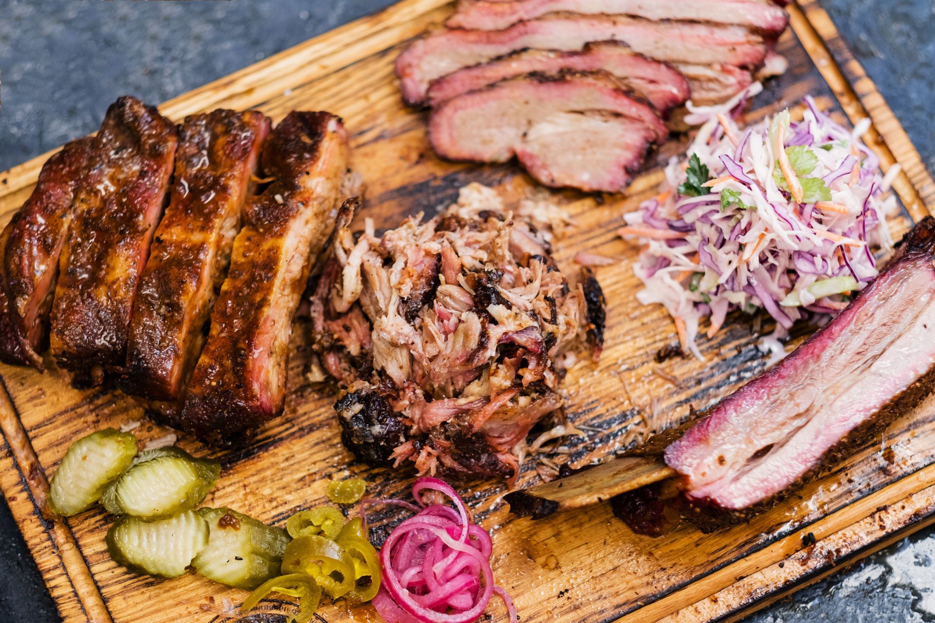 A wooden serving board features BBQ ribs, pulled pork, brisket slices, coleslaw, pickles, jalapeños, and pickled onions.