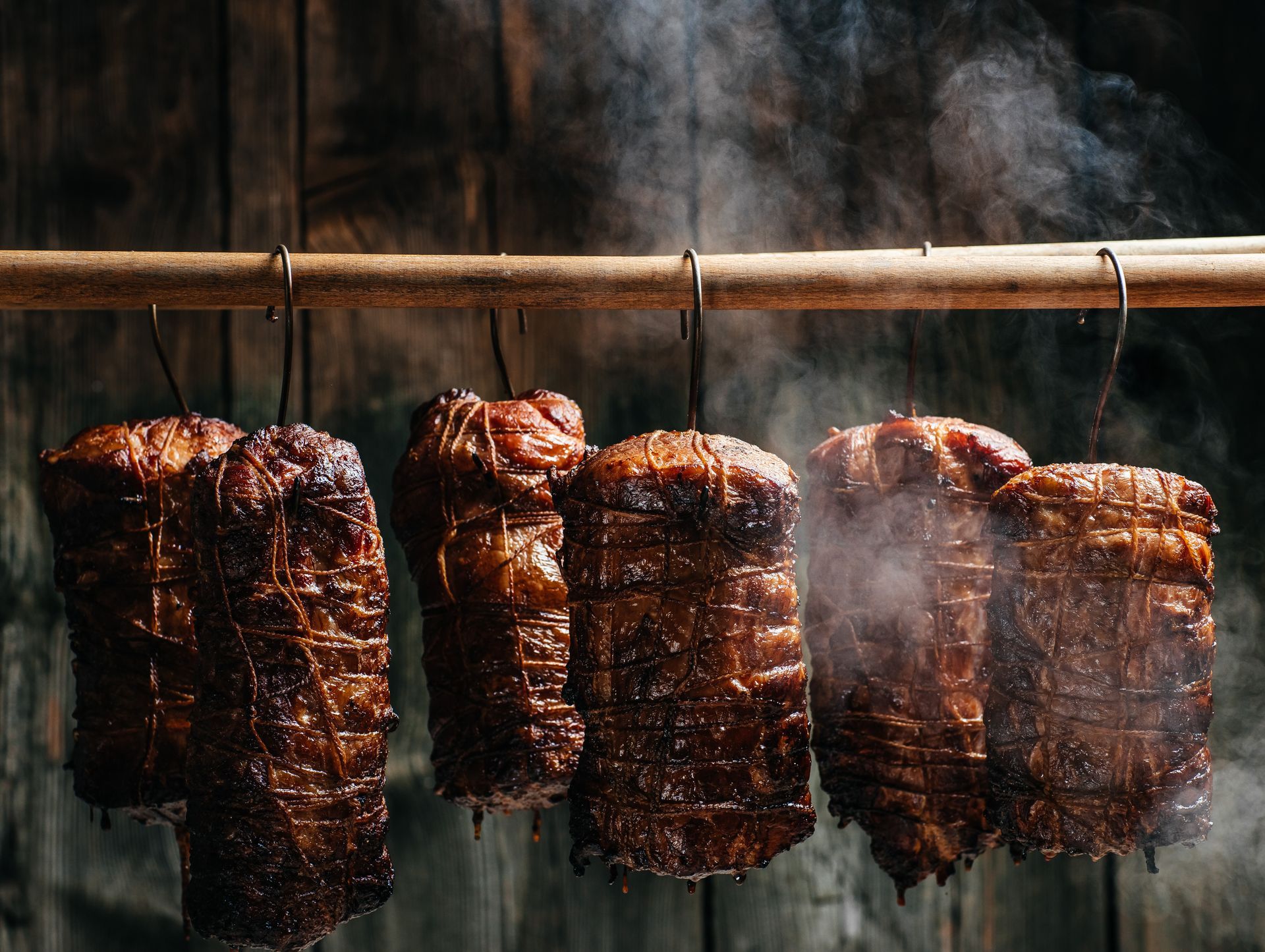 Several tied, smoked meat roasts hang from metal hooks on a wooden rod, surrounded by wisps of smoke against a dark wall.