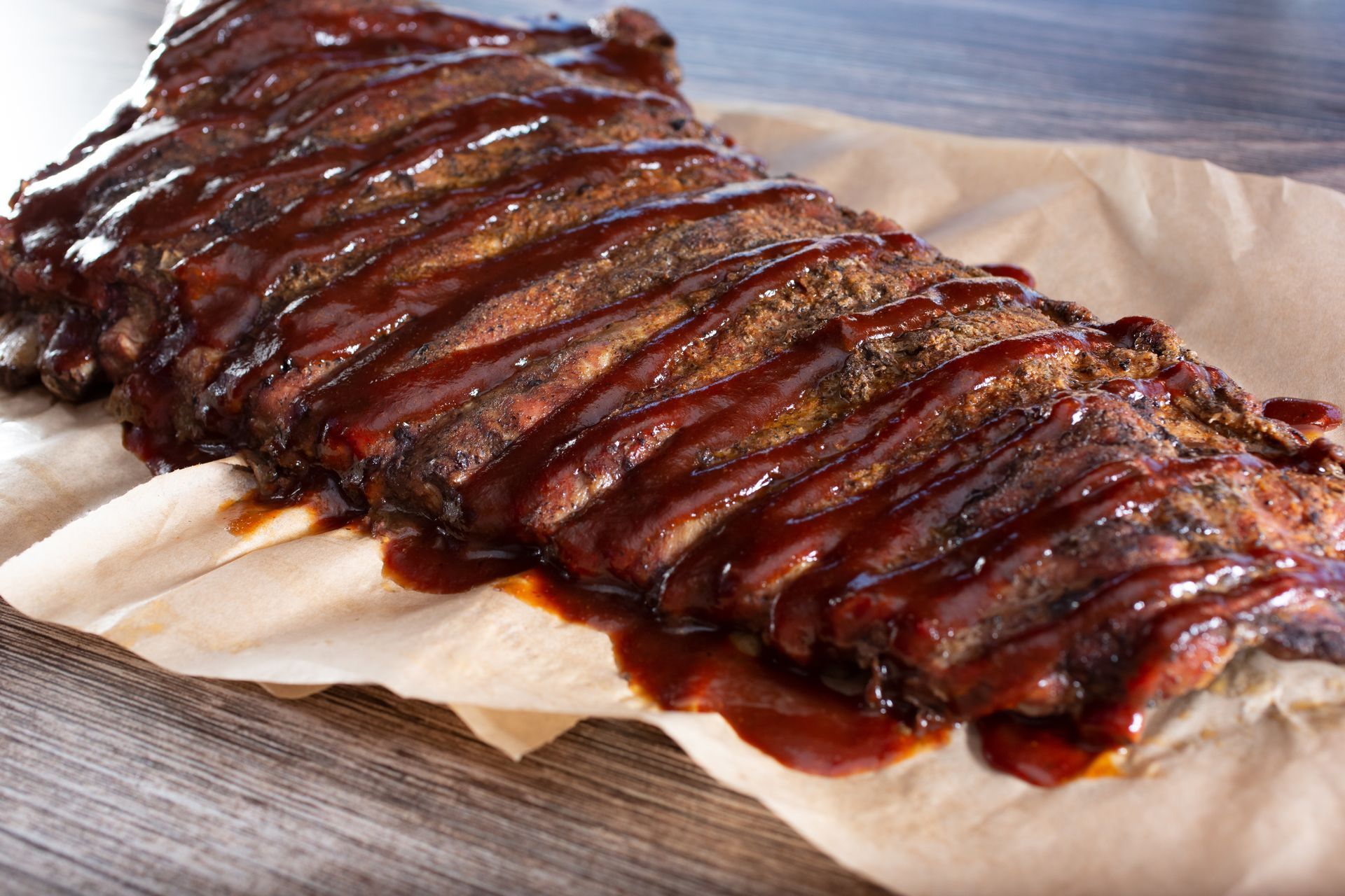 A rack of slow-cooked ribs coated in dark barbecue sauce, resting on parchment paper on a rustic wooden table.
