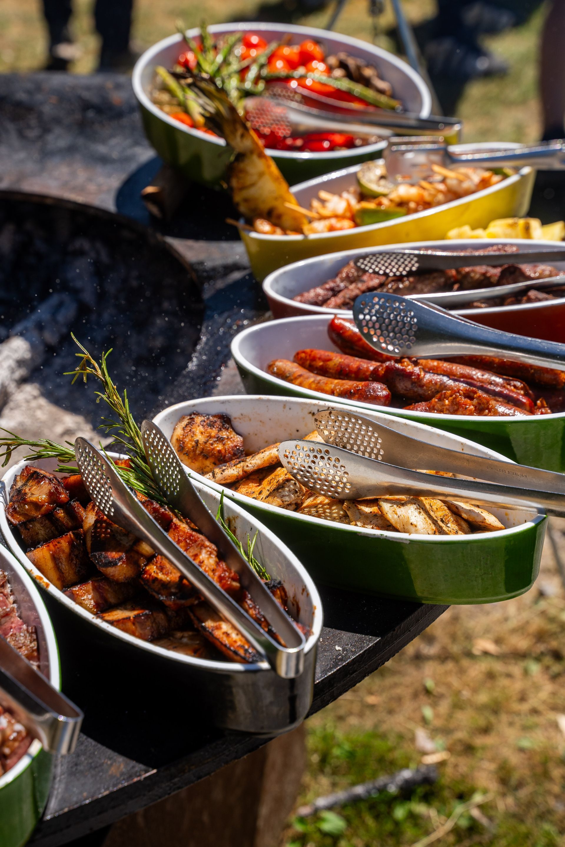 A line of colorful oval serving dishes filled with grilled meats, sausages, and vegetables on an outdoor buffet table.