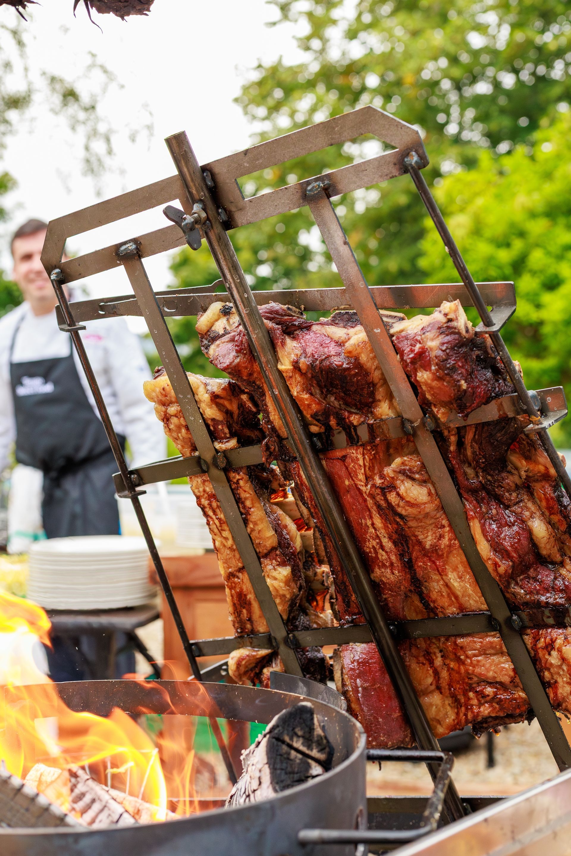 Large slabs of meat cooking in a metal rack over an open outdoor fire, with a person standing in the background.