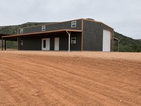 Rustic metal building on a gravel lot with a porch, white door, and wooded hills in the background