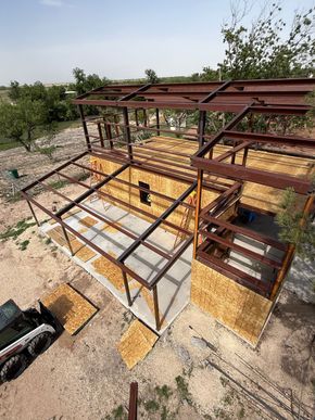 Partially built steel frame structure with rust-red beams and concrete foundation on a gravel lot