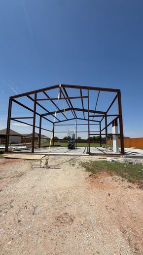 Steel building frame under construction on a gravel lot beneath a clear blue sky.