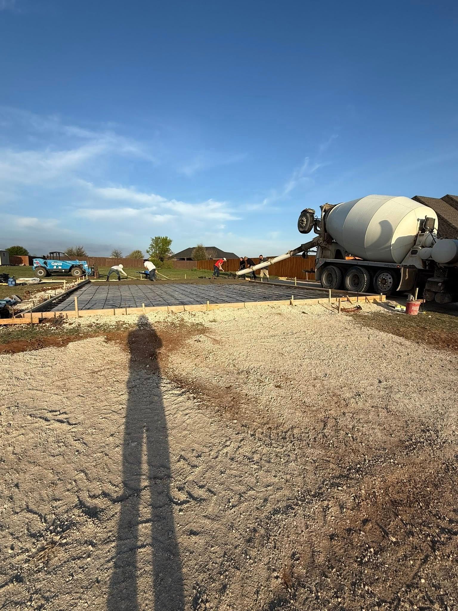 Construction site with a concrete mixer truck, gravel, and a long shadow in the foreground under a clear blue sky