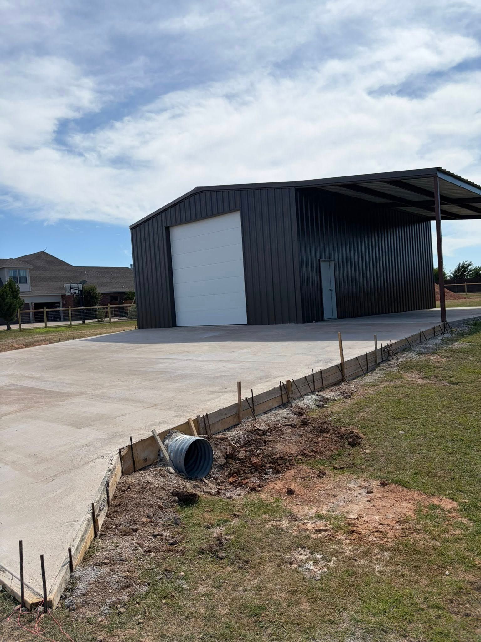 Dark metal workshop building with large white door beside a concrete driveway under a cloudy sky