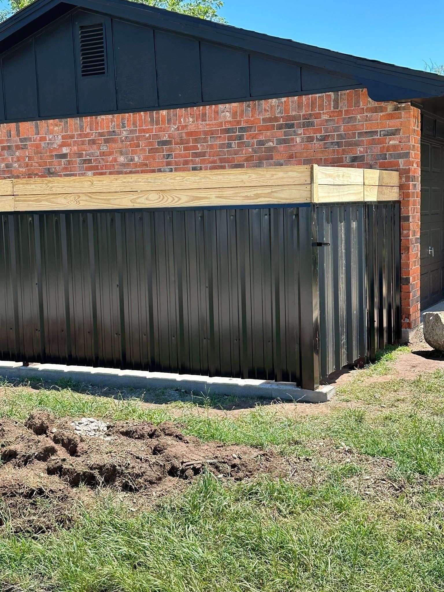 Backyard shed with dark metal walls, brick base, and a black roof beside grass and dirt.