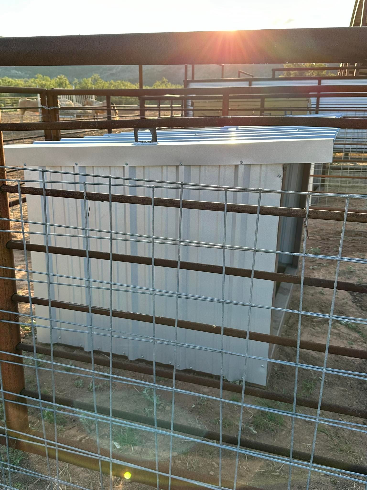 White metal container behind a wire fence in a sunlit outdoor yard with hills in the background