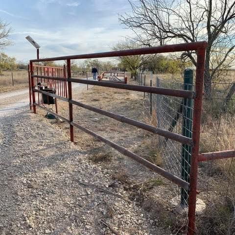 Rust-colored gate across a gravel path at a rural entrance, with fence posts and dry brush nearby.