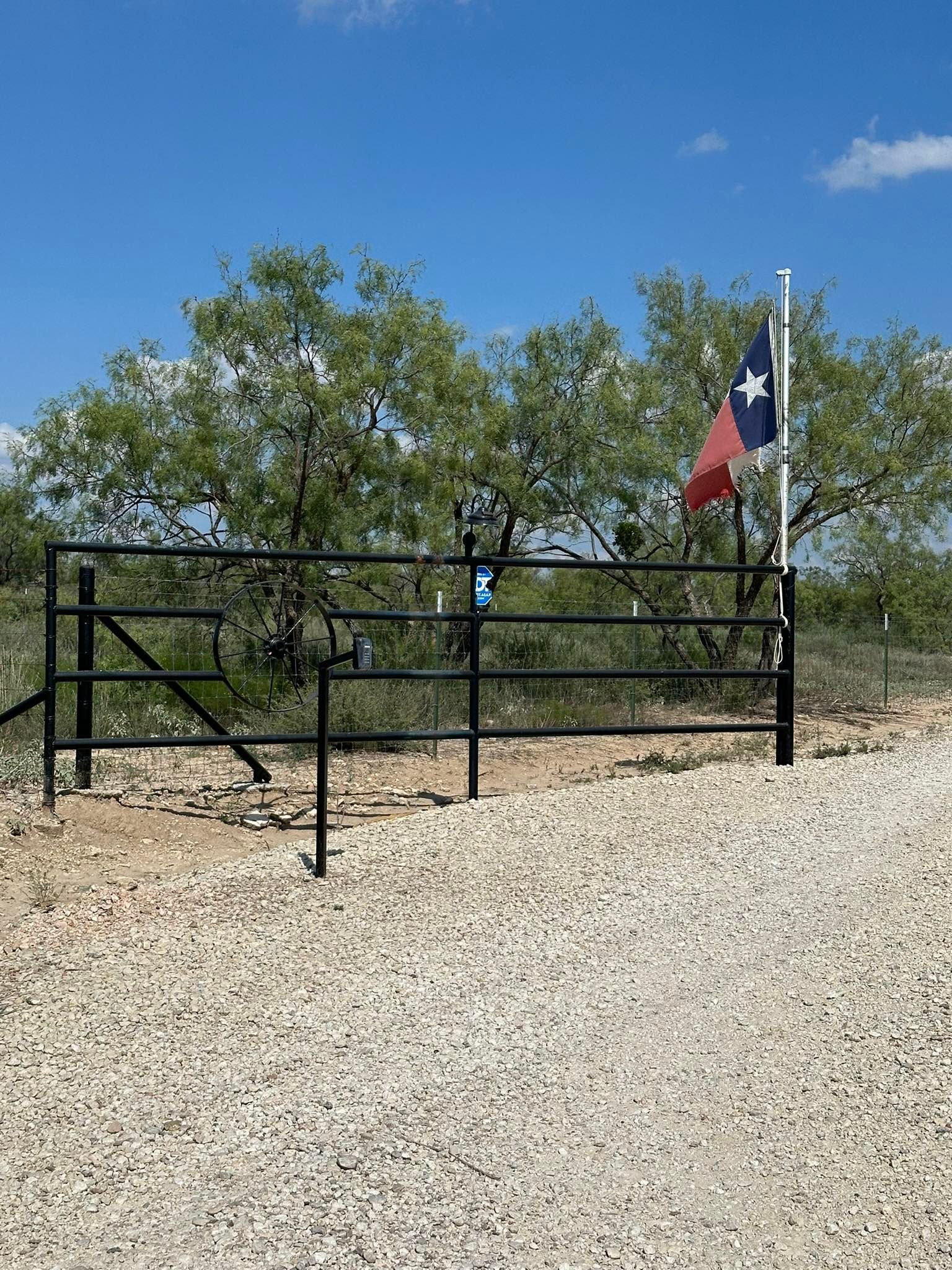 Black metal gate with Texas flag on a gravel road in a dry, brushy landscape under a clear blue sky