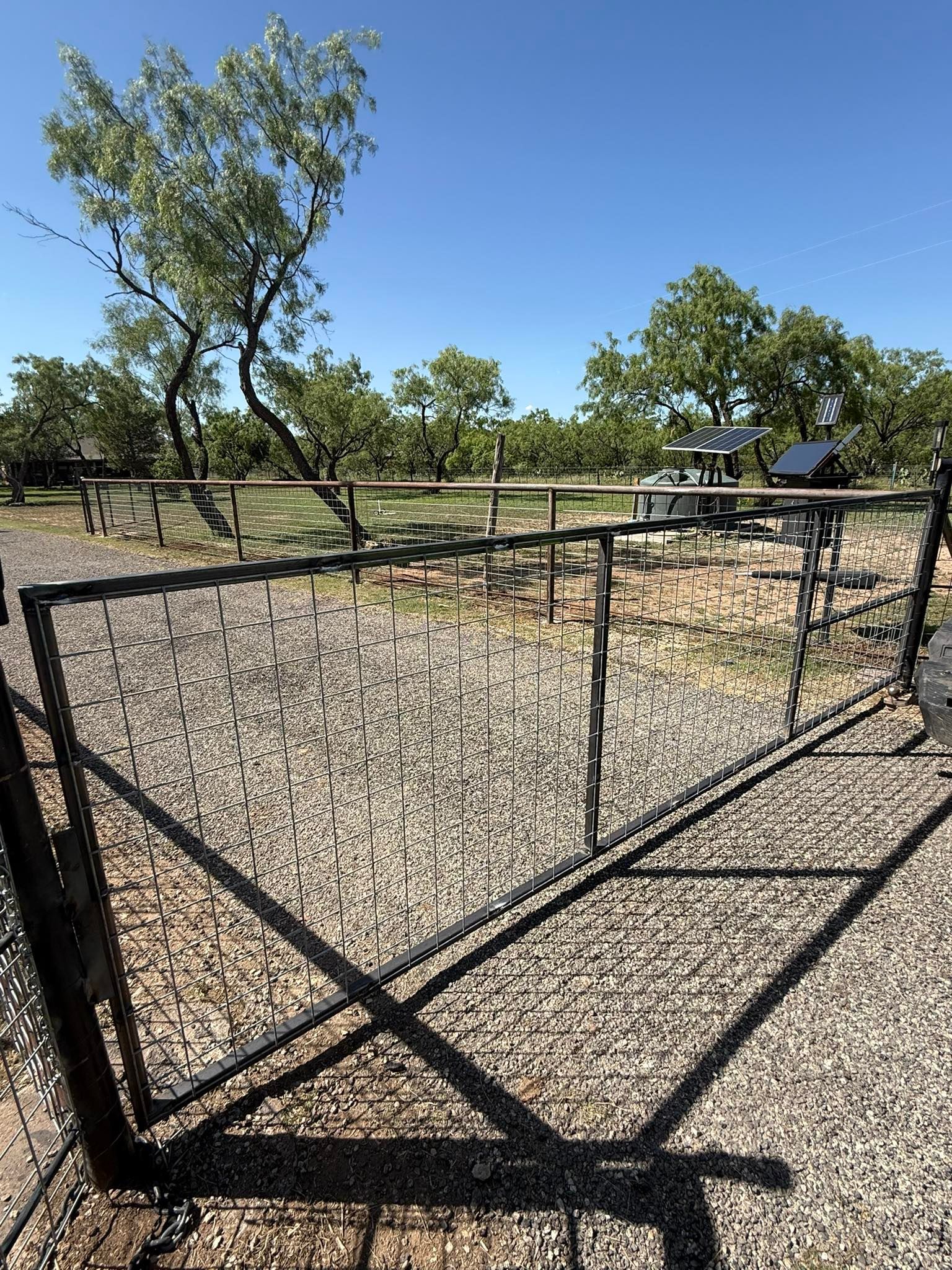 Black metal gate in a gravel yard with dry trees and a blue sky behind it
