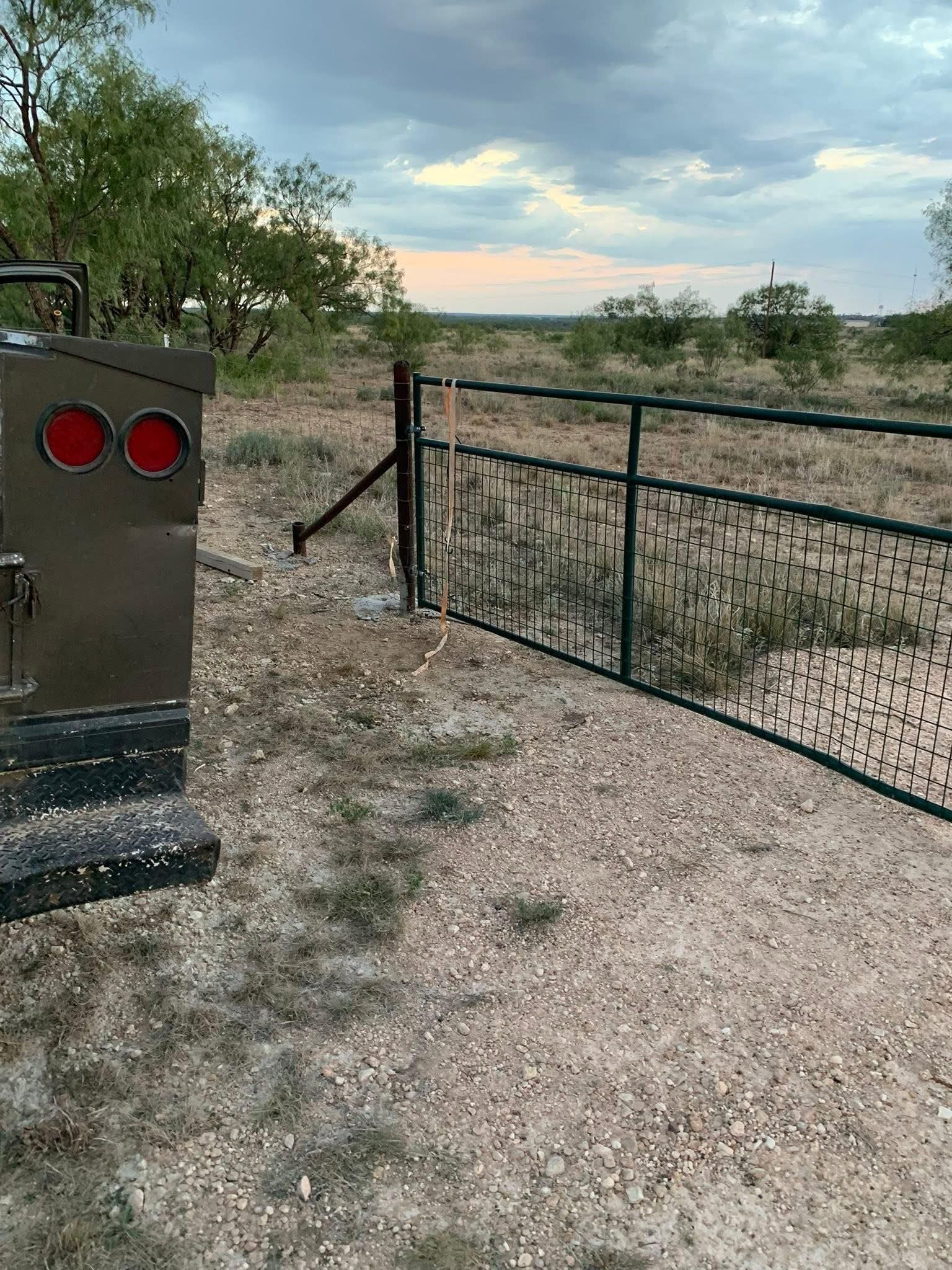 Open gate in a gravel driveway with trees and a sunset sky beyond.