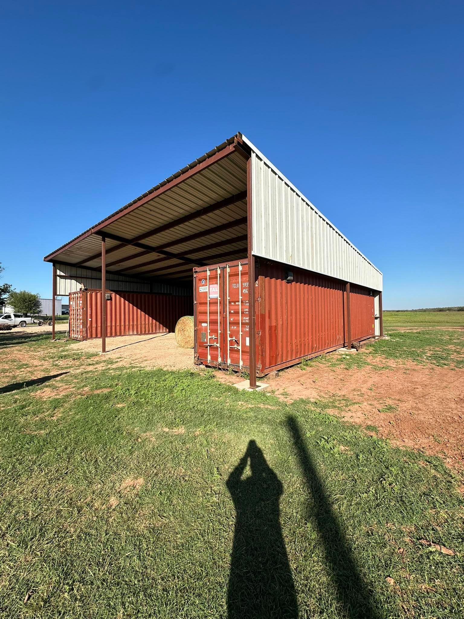 Red barn-like shed with a metal roof in a grassy field under a clear blue sky.