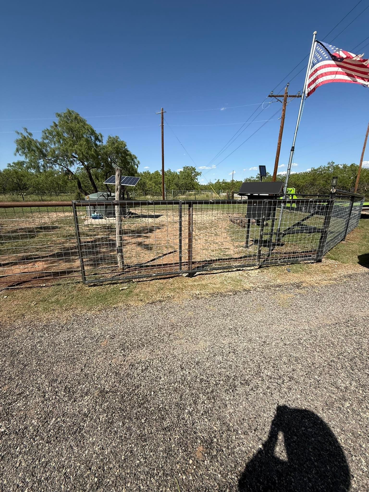 Gravel yard with chain-link fence, trees, and a large American flag on a pole under a clear blue sky