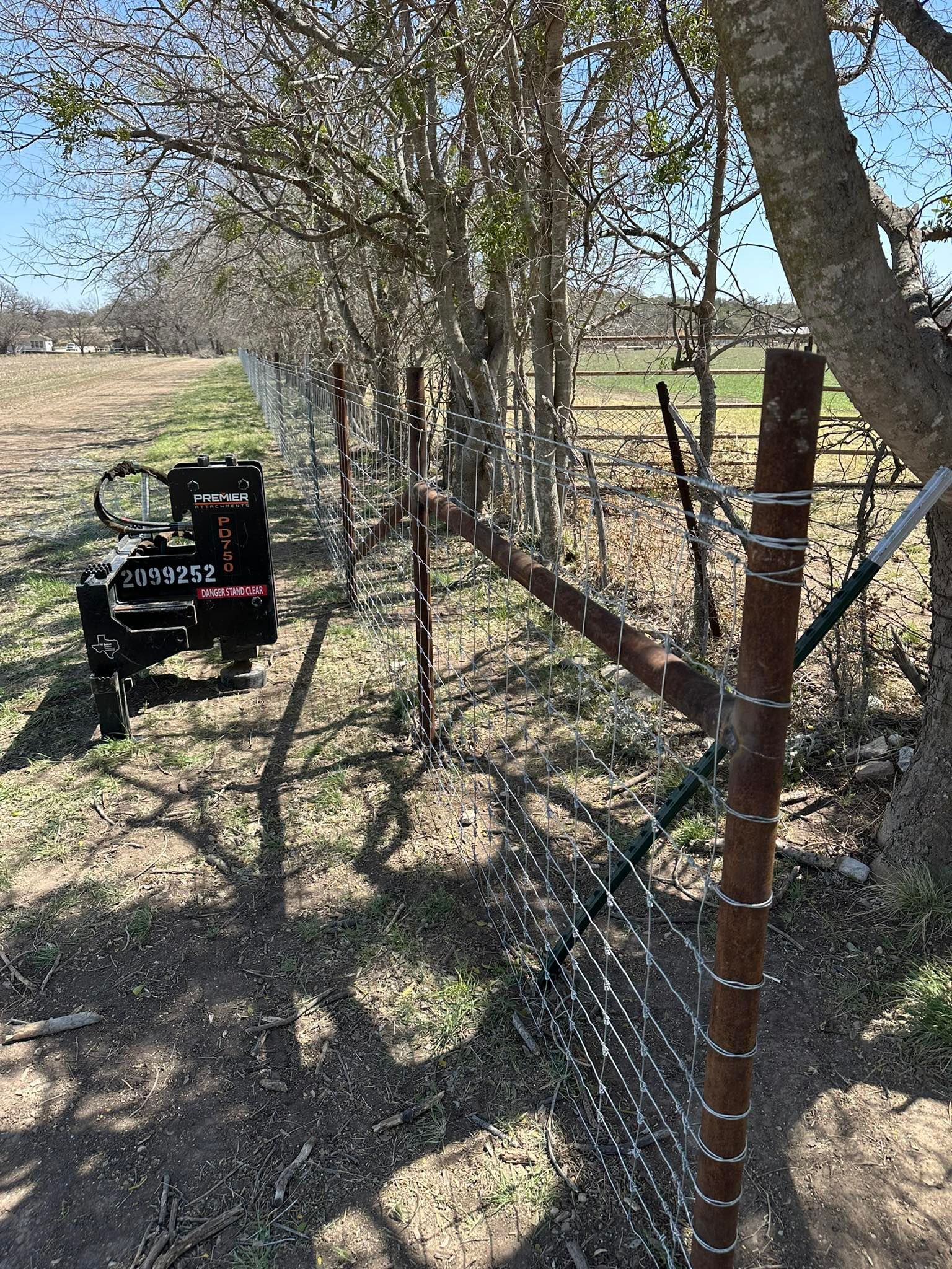 Fence-lined dirt path beside a tree in a rural field, with a small wagon on the left and a metal fence post in front.