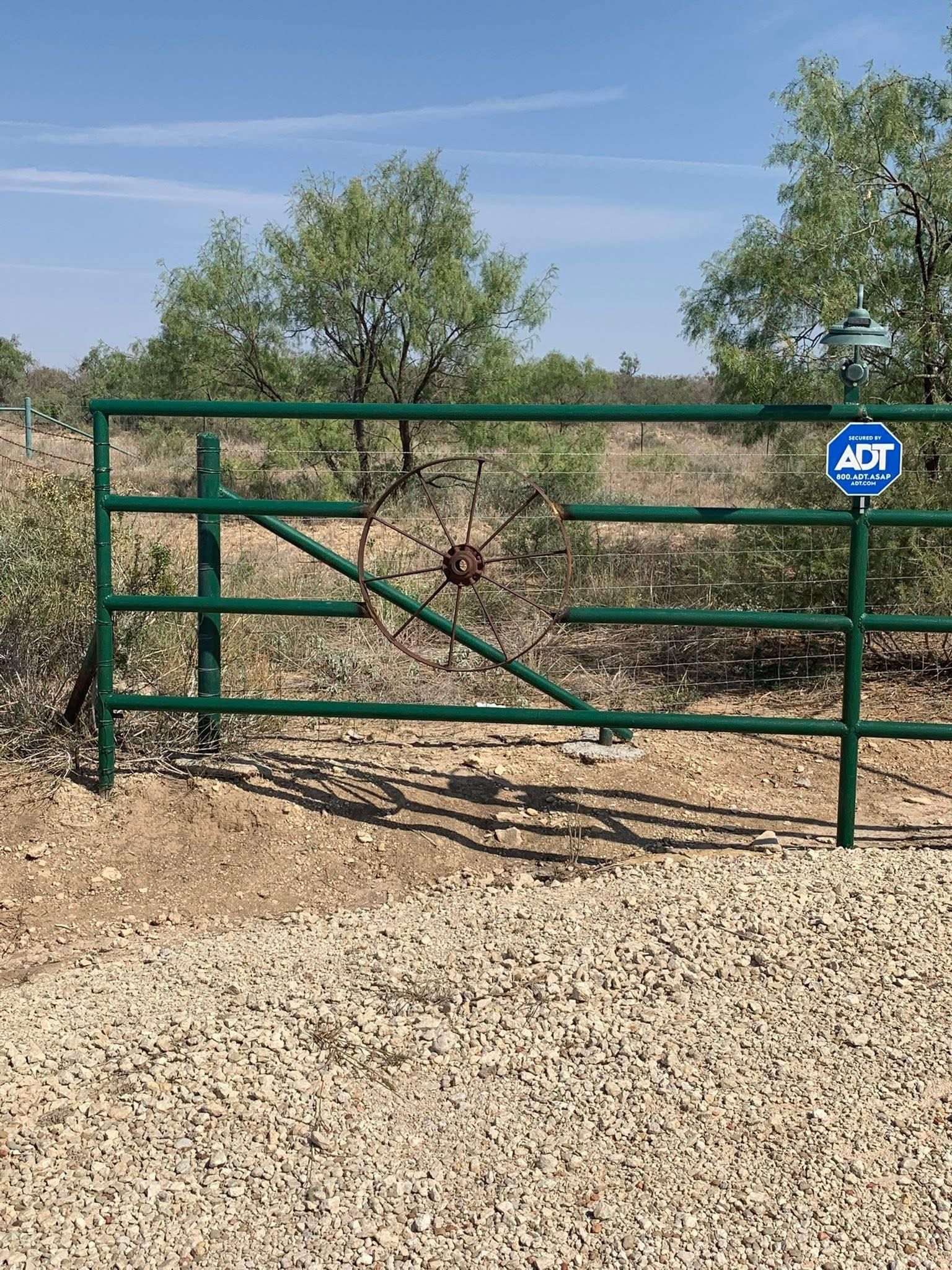 Green metal ranch gate on a gravel road in a dry rural landscape with an ADT sign.