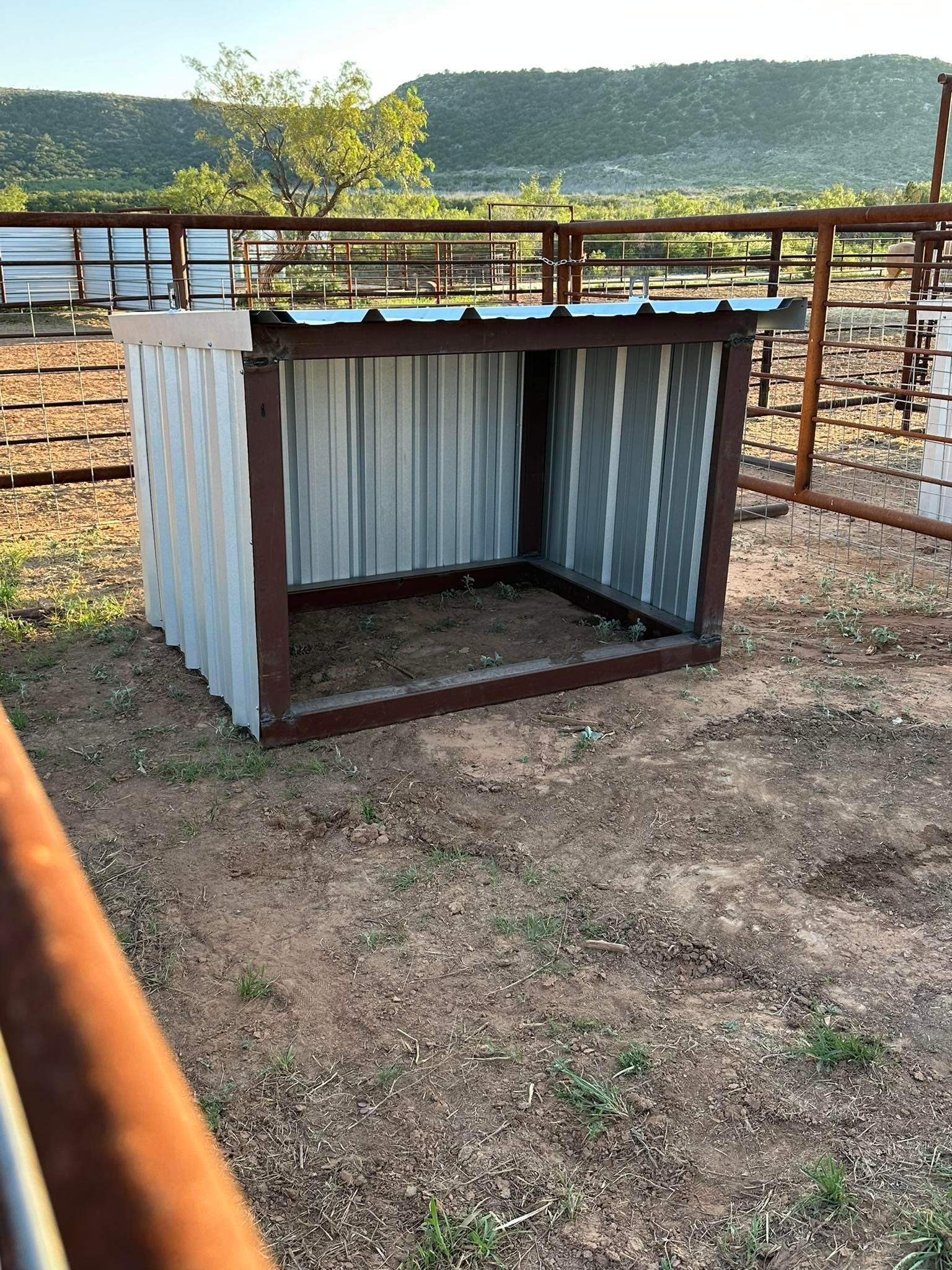 Small corrugated metal shelter in a dusty livestock pen with fences and hills in the background