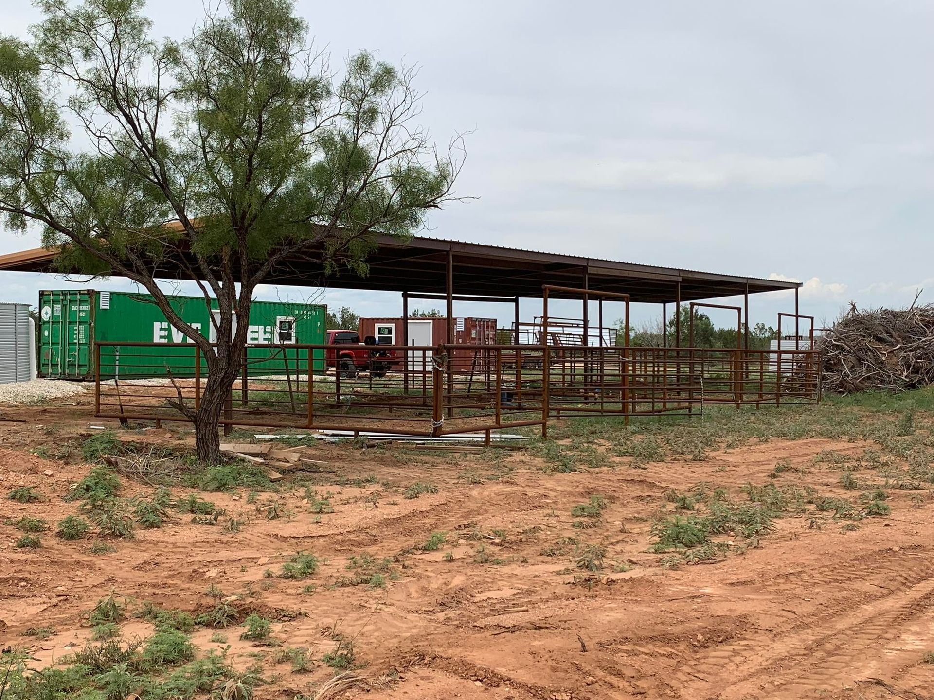 Rural livestock pen with metal fencing, a shed roof, and a green building on dry, reddish ground