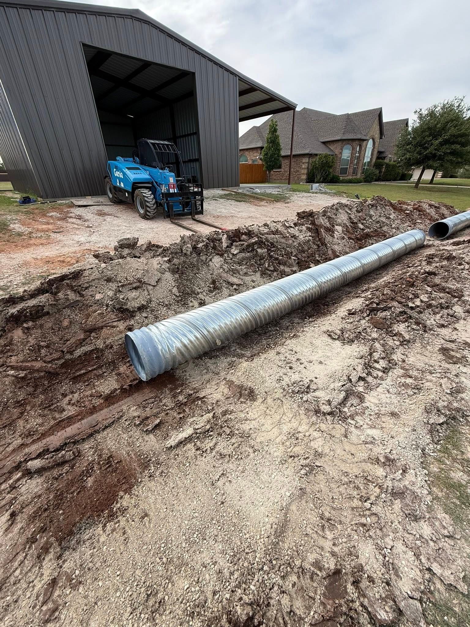 Construction site with a blue tractor by a shed and a long metal pipe in a dirt trench