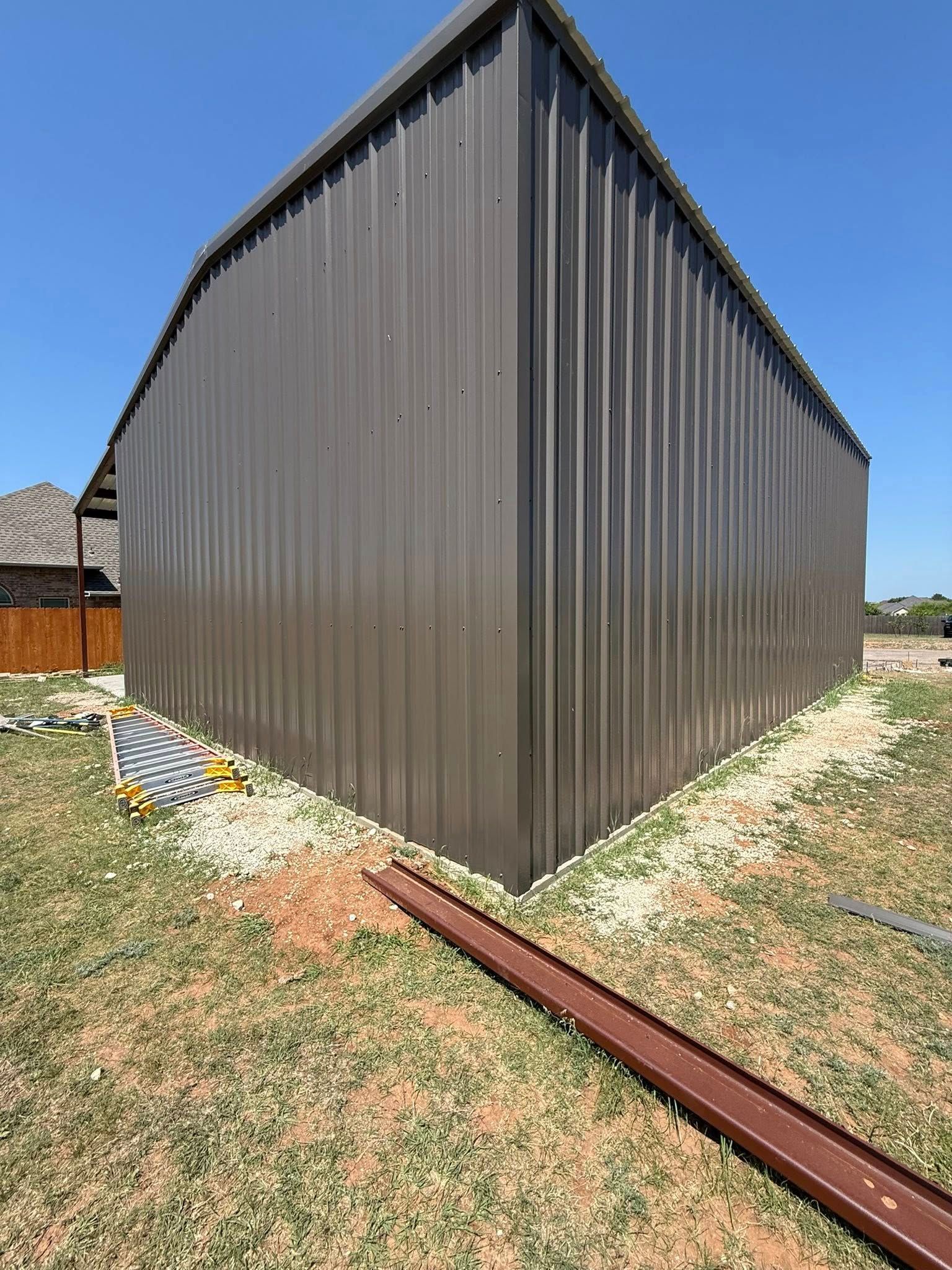 Dark gray corrugated metal shed with a sloped roof on a gravel yard under a blue sky