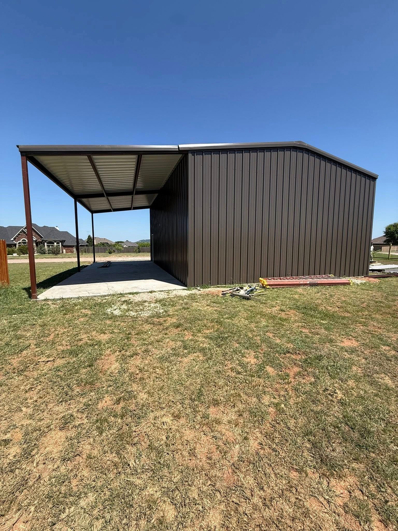 Small dark metal shed with a covered side area on a grassy rural lot under a clear blue sky.