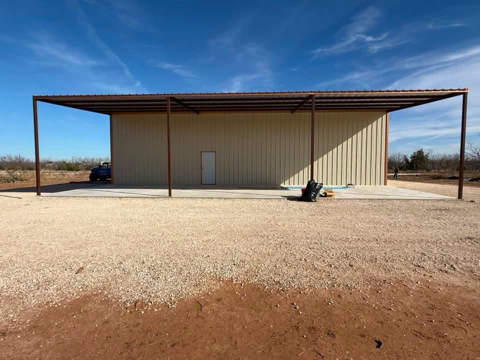 Large beige metal building with a covered open-sided awning on a gravel lot under a clear blue sky