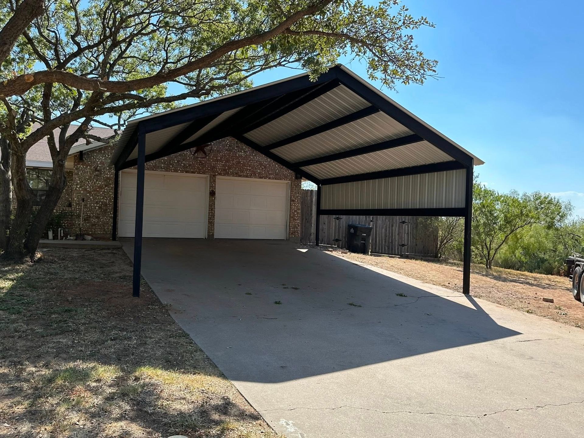 Covered driveway carport with two garage doors, black metal posts, and a sloped roof beside a brick house.