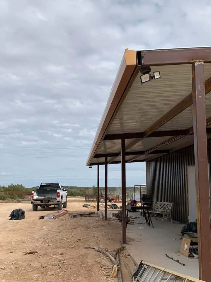 Rural worksite with metal-roofed shelter, pickup truck, tools, and dirt driveway under cloudy sky
