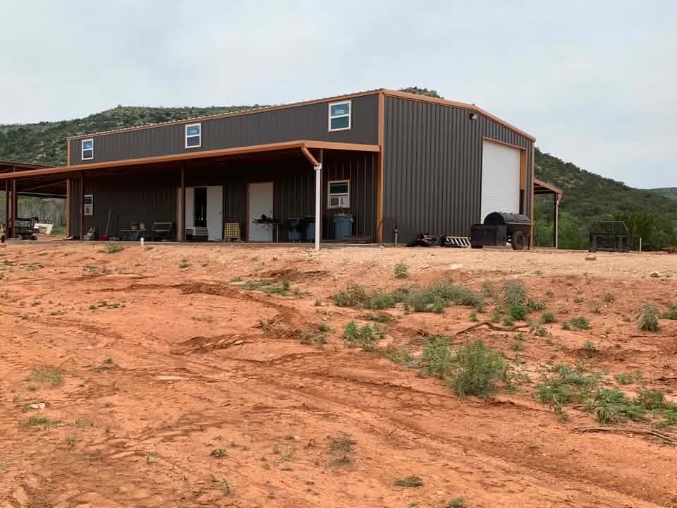 Rustic metal house on a red dirt lot with a covered porch and green hillside backdrop