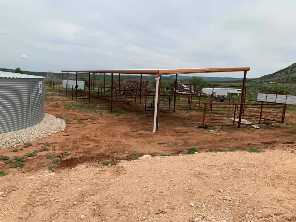 Open-sided metal shed on red dirt near a water tank and hillside under cloudy sky