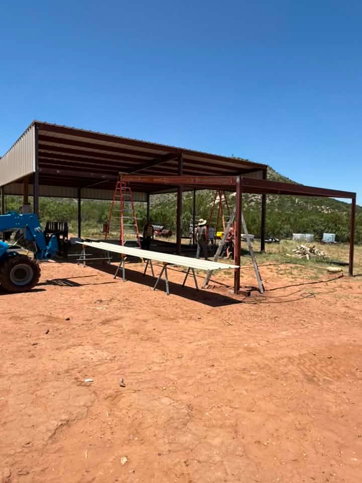 Open-sided metal shed under construction on a dirt lot, with a blue tractor and workers nearby.