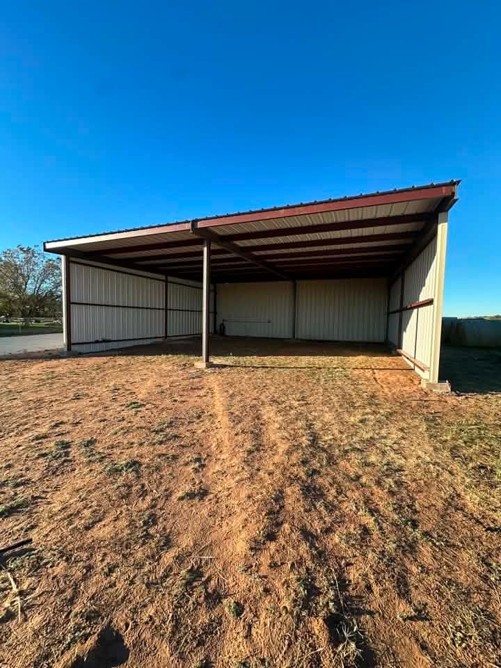 Open-sided metal shed with a red roof on a dirt lot under a clear blue sky