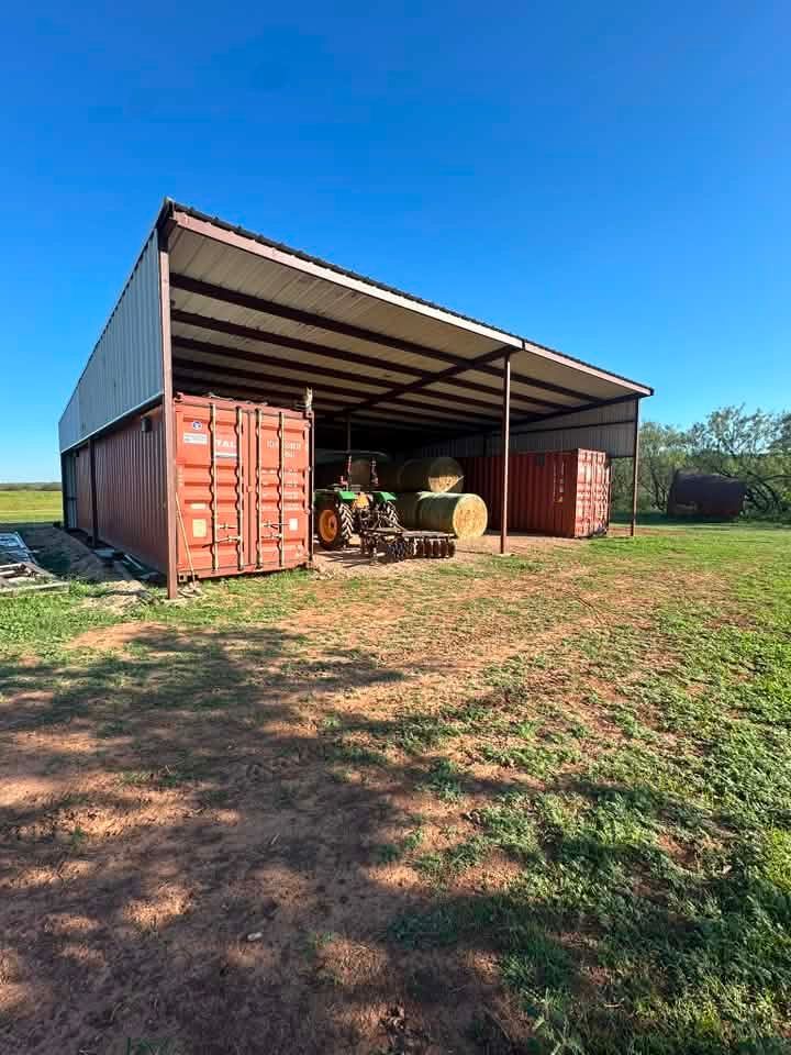 Open-sided barn with shipping containers and farm equipment in a grassy field under a blue sky