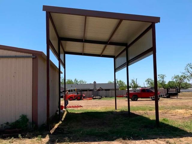 Open-sided metal carport beside a beige building, with red trucks parked in the background.