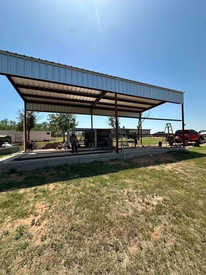 Open-sided pavilion with a metal roof, benches, and a red truck beside a grassy field under a clear blue sky