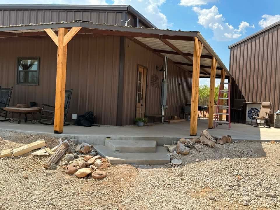 Brown building with wooden porch posts, concrete steps, and gravel yard under a blue sky
