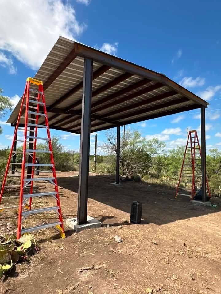 Partially built metal-roofed shelter with ladders on both sides in a dirt outdoor area