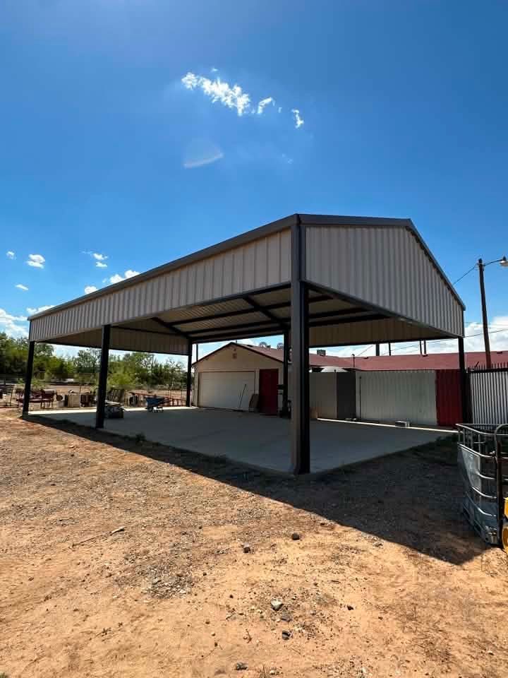 Large metal carport beside a small building on a sunny, dirt lot under a blue sky.