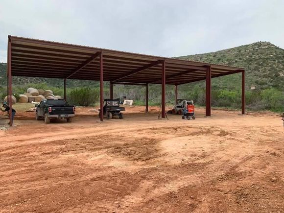 Open-sided metal shed with parked trucks and a small tractor on a dirt lot near rocky hills.