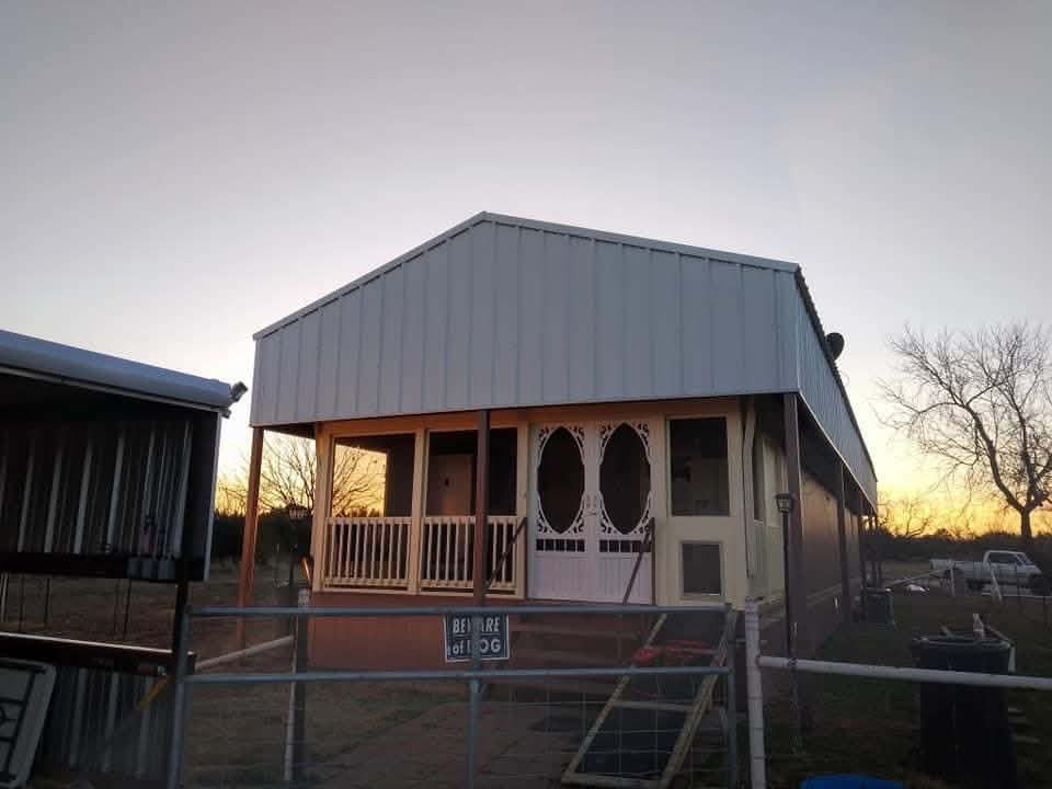 Small porch-front house with metal siding at sunset, viewed from the street behind a chain-link fence.