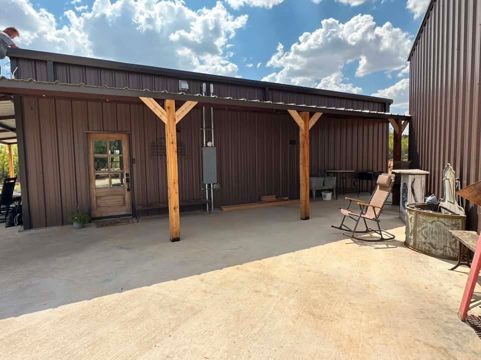 Brown metal building with a covered patio, wooden posts, chairs, and a sunny concrete courtyard