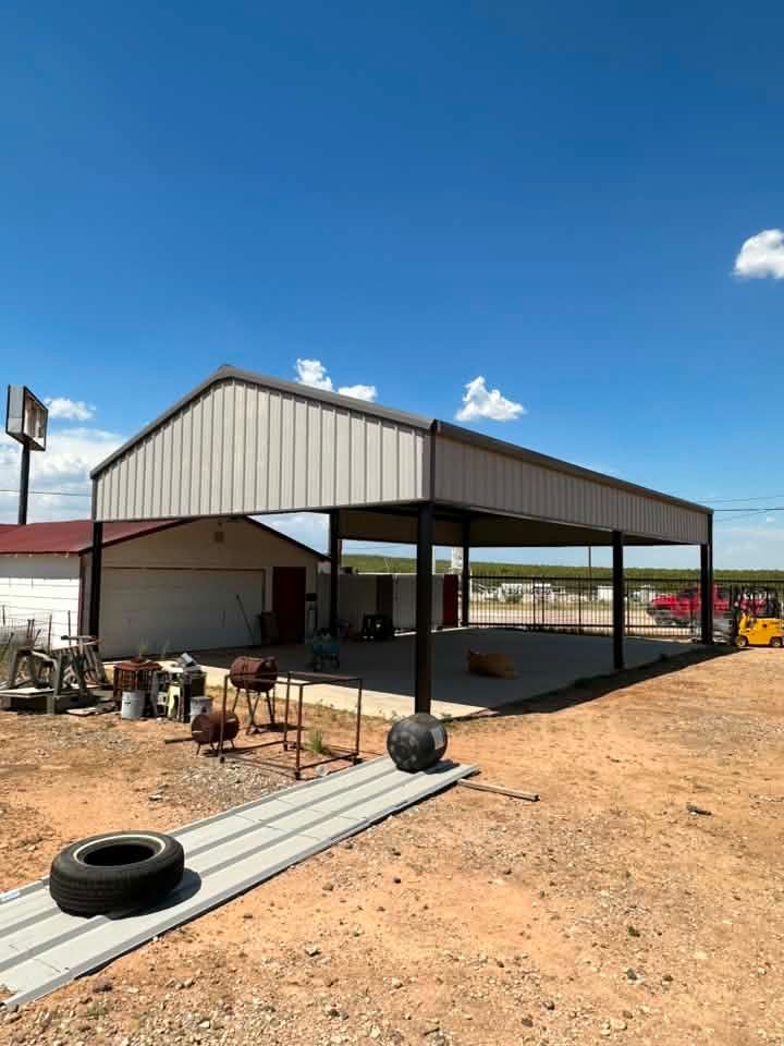 Open-sided metal barn with work equipment on a dirt lot under a blue sky