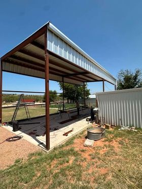 Open-sided metal shed with a corrugated roof on a dirt lot beside a white fence