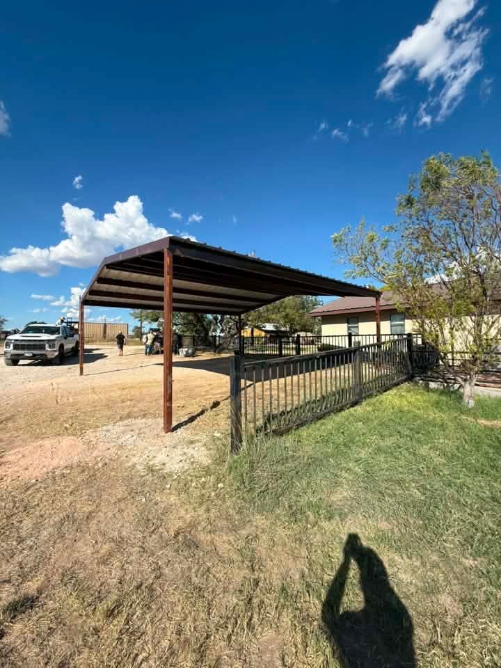 Open metal carport in a dusty yard under a blue sky, with a fence and parked truck nearby