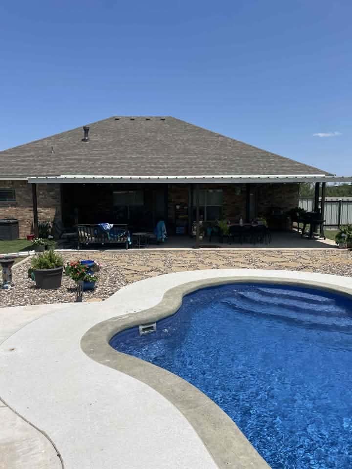 Backyard pool beside a covered patio and stone house under a clear blue sky