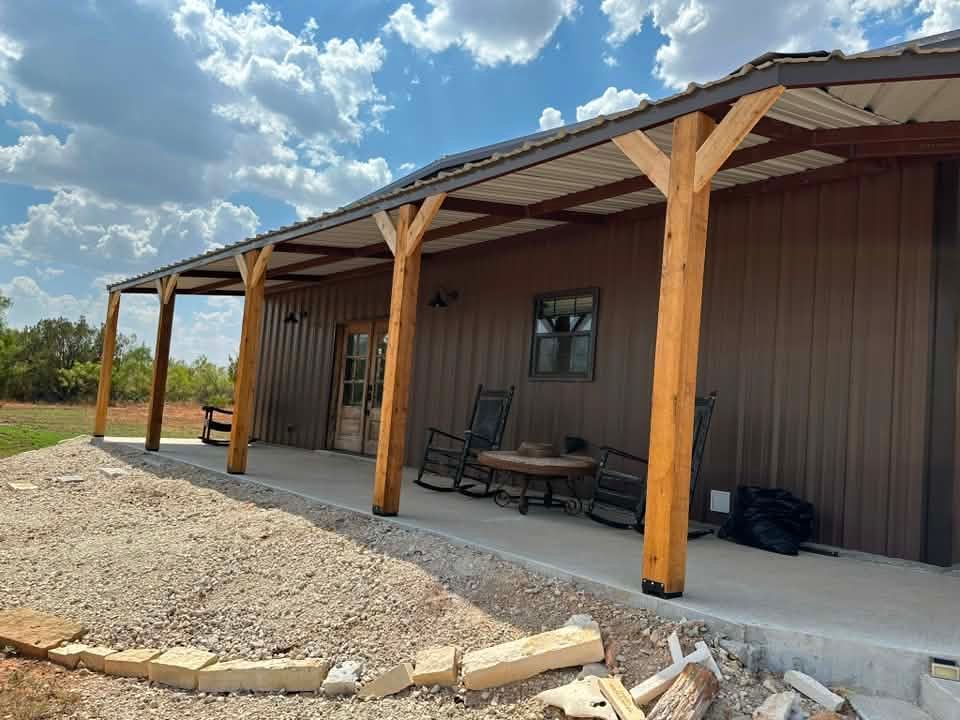Covered porch with wooden posts on a brown metal building, gravel foreground, and blue cloudy sky.