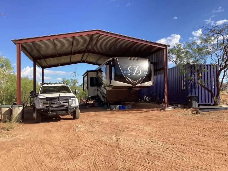 Rustic yard with a carport sheltering a white truck and a large trailer beside stacked shipping containers.